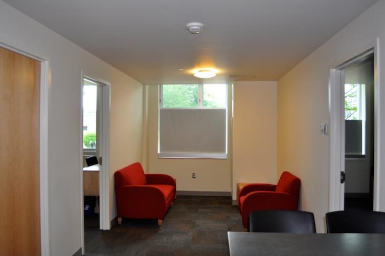 Living room with red couch and red chair. The blindes are pulled up halfway (bottom of window is covered) to let in light while providing privacy from the street. The floor is carpeted.